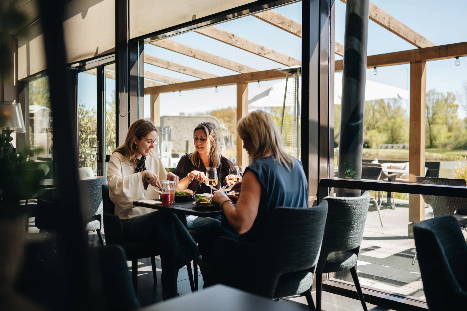 Drie vrouwen genieten van een lunch met witte wijn aan een tafel bij de grote glazen pui binnen in het restaurant, met direct uitzicht op het zonnige terras.
